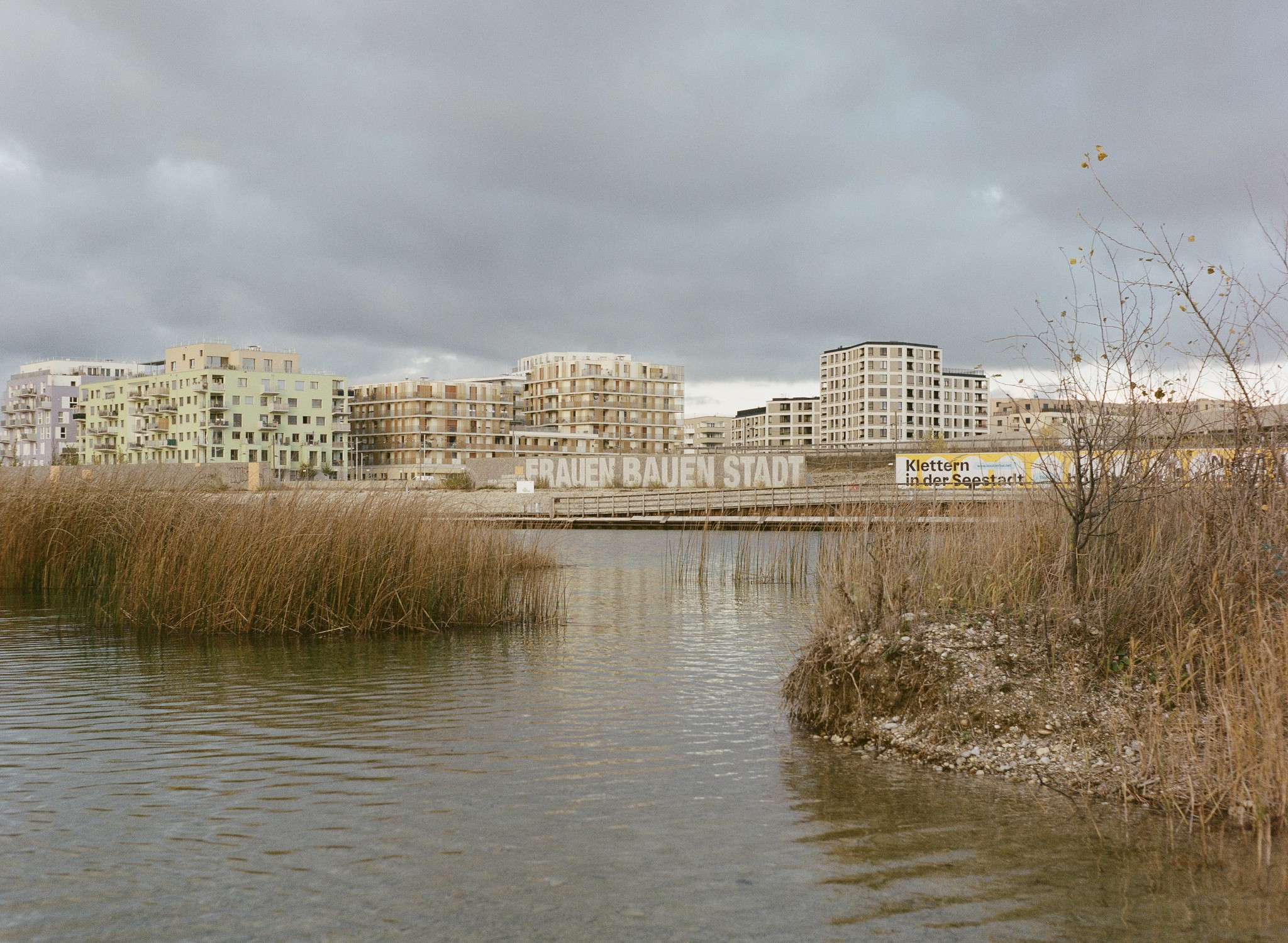 Blick auf die erweiterte Seestadt am anderen Ende des Sees. Auf der Hauswand steht in großen Lettern "Frauen bauen Stadt" geschrieben.