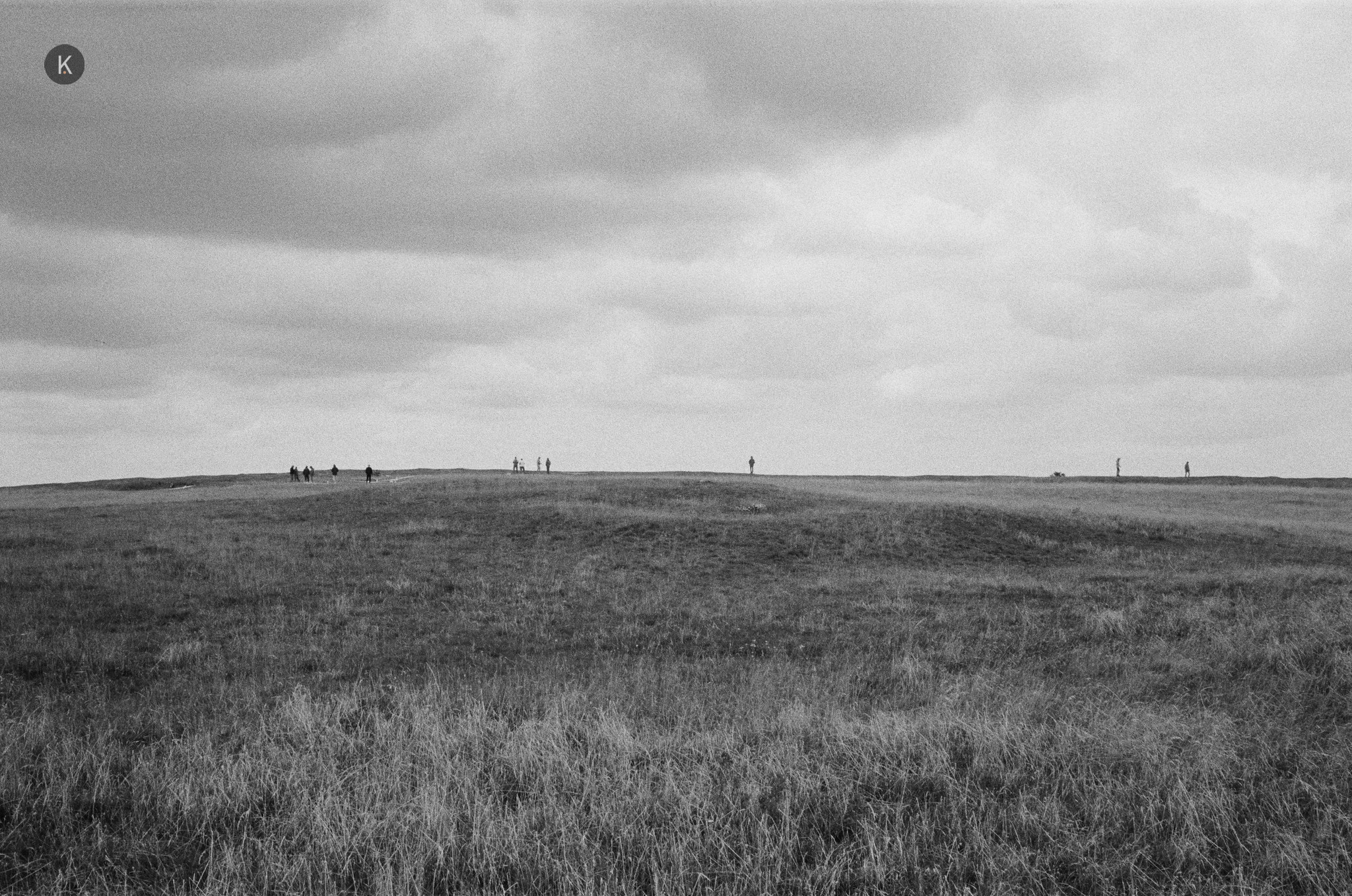 Schwarz-weiß-Foto einer weiten, leeren Wiesenlandschaft unter bewölktem Himmel, mit kleinen, verstreuten Menschen in der Ferne am Horizont.