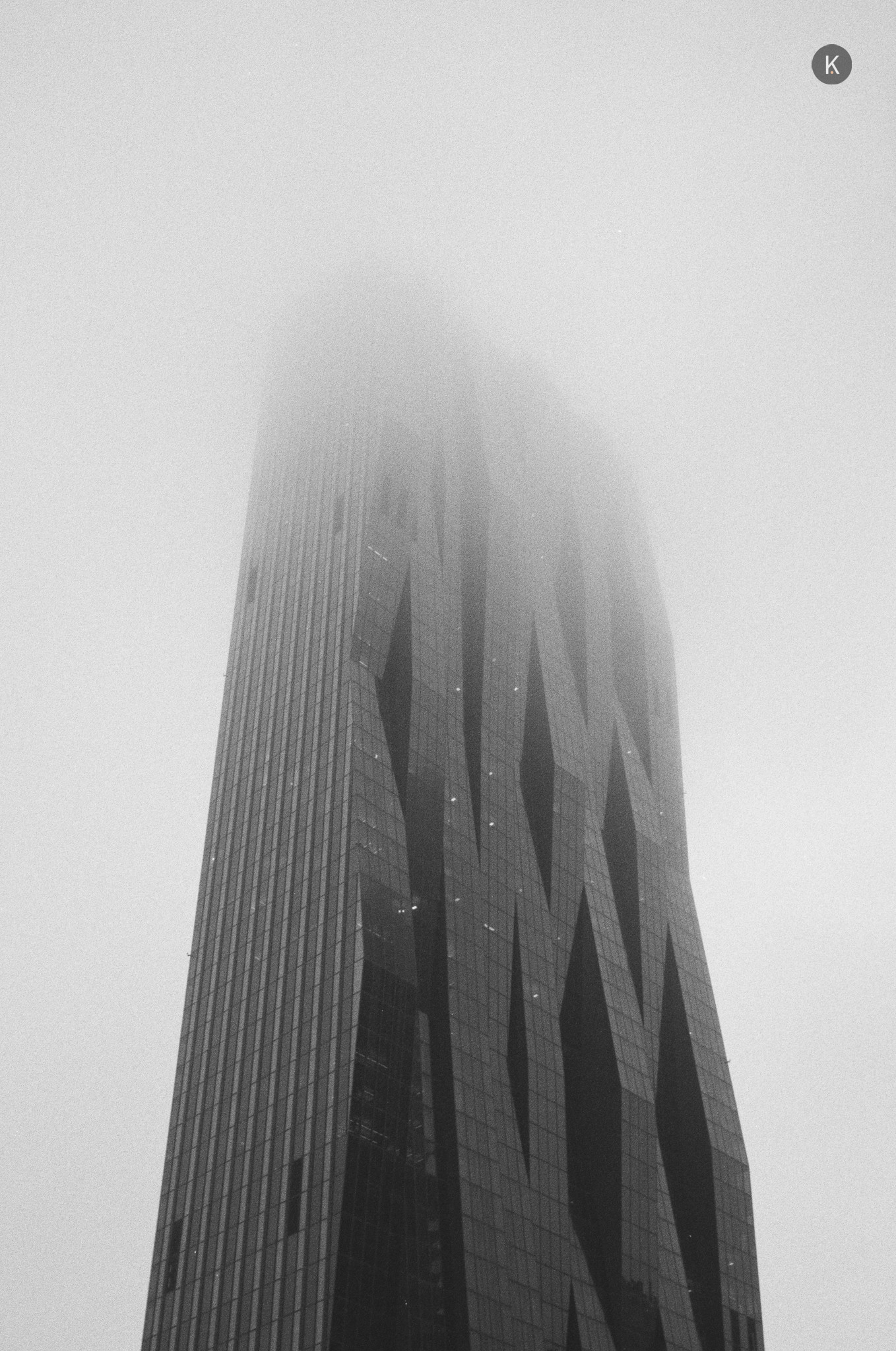 Ein schwarzweißes Foto eines hohen, modernen Wolkenkratzers, dessen Spitze im dichten Nebel verschwindet; die Fassade hat dunkle, diagonale Muster und das Gebäude wirkt von unten fotografiert.