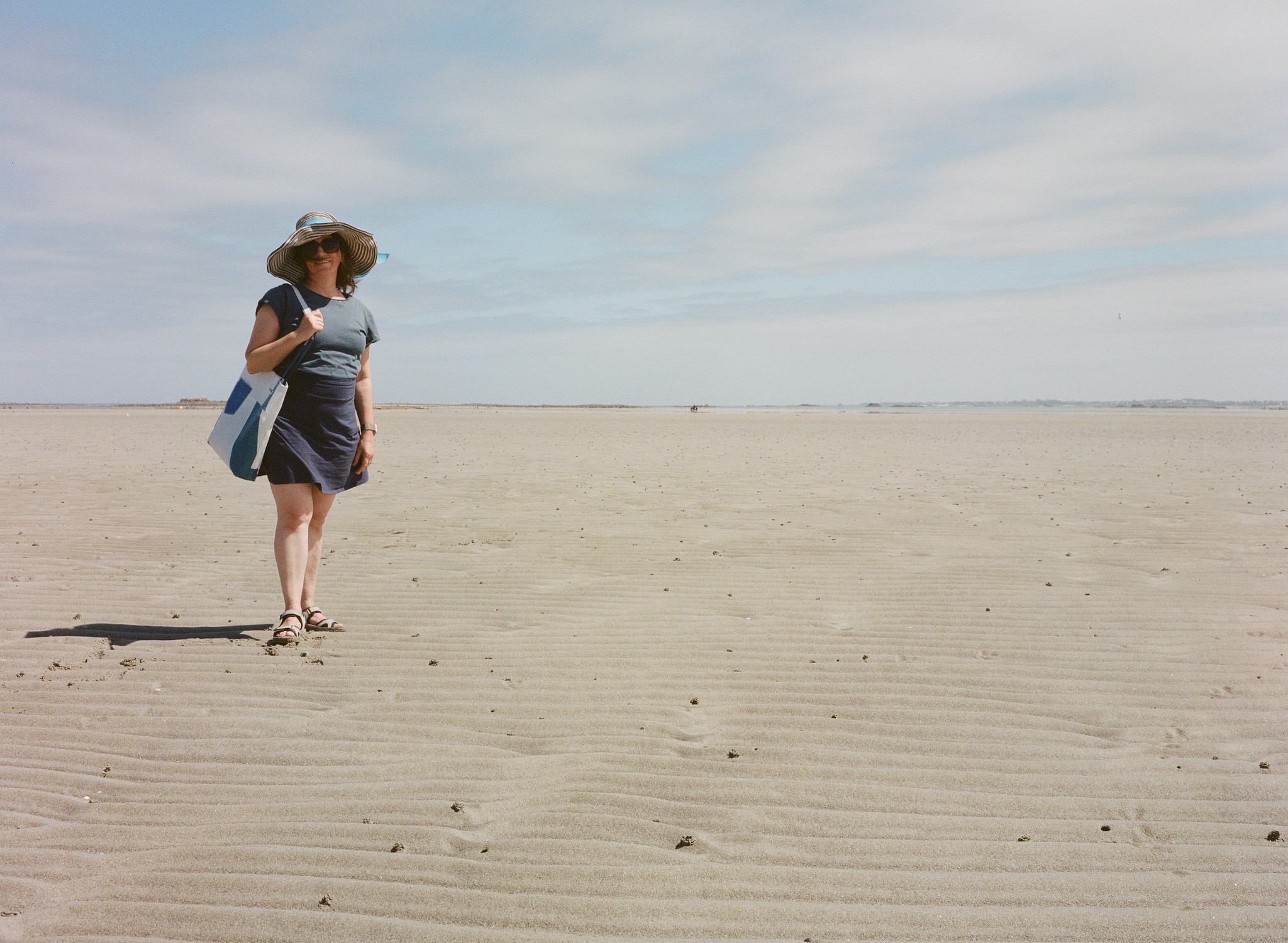 Frau mit Sonnenhut und Stofftasche steht auf einem weitläufigen, menschenleeren Sandstrand unter leicht bewölktem Himmel.