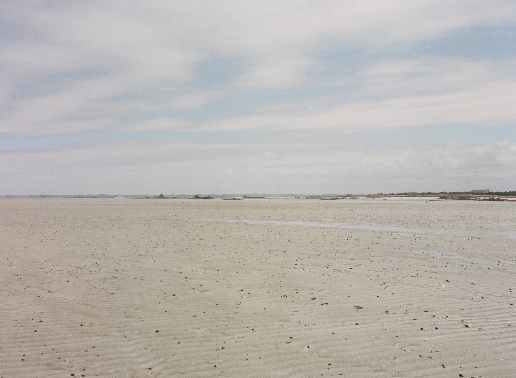 Weite, bei Ebbe freiliegende Sandfläche mit flachen Rillen unter einem bewölkten Himmel; am Horizont liegen niedrige Dünen und eine schmale Küstenlinie.
