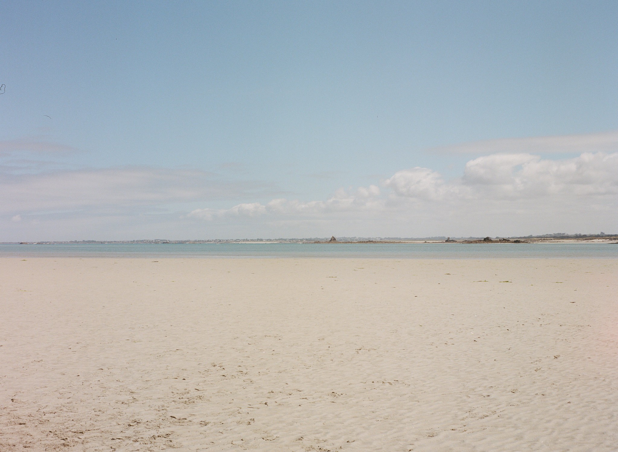 Weitläufiger, fast menschenleerer Sandstrand mit ruhigem, türkisblauem Meer unter einem hellblauen Himmel mit wenigen Wolken.